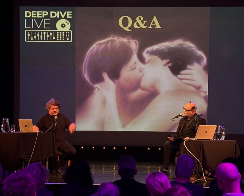 Ed Buller and Simon Price on stage during the Q&A segment, with a screen behind them showing an embracing couple and the words 'Deep Dive Live Q&A'
