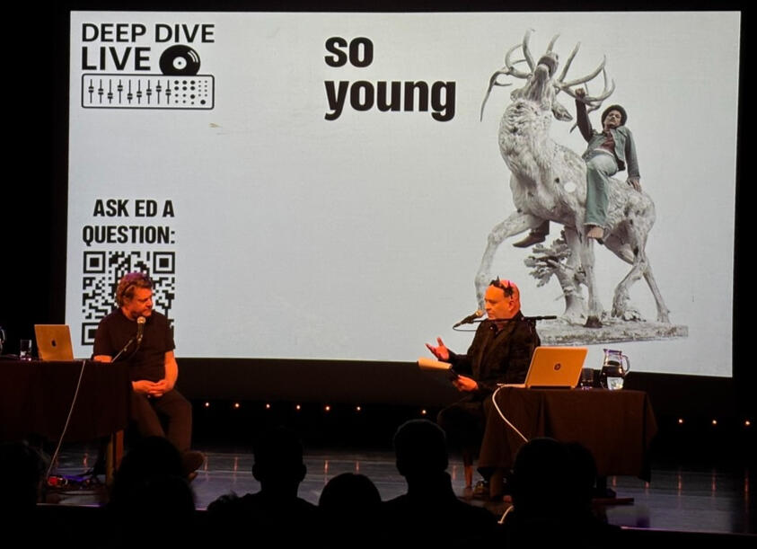 Ed Buller and interviewer Simon Price seated on stage with a black-and-white photo of So Young single artwork displayed on the screen behind them.